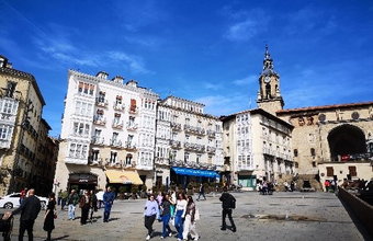 Heller Platz mit alten Gebäuden, Balkonen und einem Kirchturm im Hintergrund. Menschen schlendern über den Platz bei blauem Himmel.
