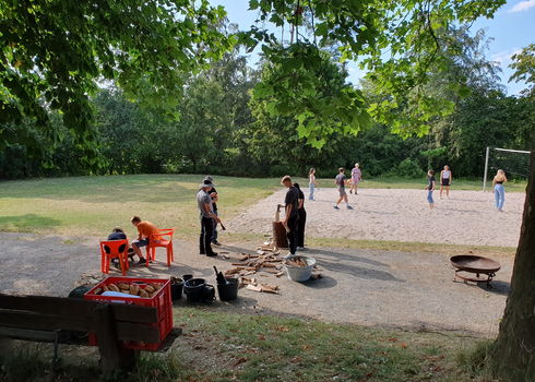 Im Vordergrund hacken Schüler Holz und bereiten Feuerholz vor. Dahinter spielen andere Jugendliche Volleyball im Sandfeld. Die Szene spielt an einem sonnigen Tag auf einer Wiese mit Bäumen.