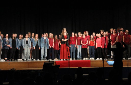 Ein großer gemischter Chor steht auf einer Bühne und singt. Die Sänger*innen tragen überwiegend rote oder jeansfarbene Kleidung. Eine Solistin in einem roten Kleid steht in der Mitte, vor der Bühne dirigiert eine Person.