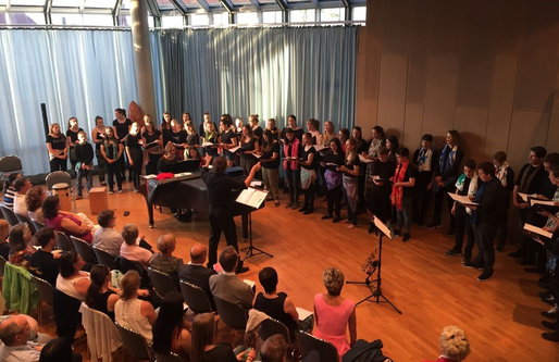 Ein großer Chor steht in einem hellen Konzertsaal mit Glasdach und singt. Eine Dirigentin leitet den Chor, begleitet von einer Pianistin am Flügel. Das Publikum sitzt in mehreren Reihen und hört aufmerksam zu.