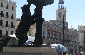 Statue eines Bären, der an einem Baum steht, auf einem Platz in der Innenstadt von Madrid. Im Hintergrund sind Gebäude und Passanten zu sehen.