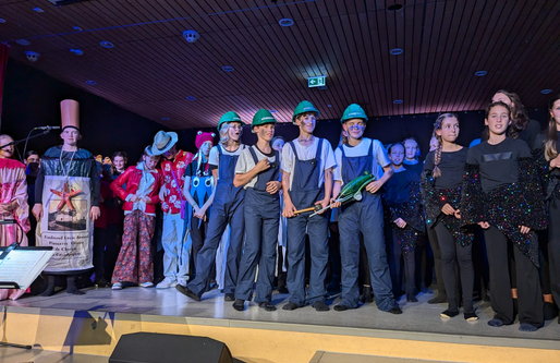 Schüler*innen stehen auf einer Bühne und tragen verschiedene Kostüme, darunter Overalls mit grünen Helmen, bunte Fantasieverkleidungen und schwarze Glitzeroutfits. Die Gruppe singt und spielt im Scheinwerferlicht.