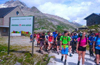 Eine große Wandergruppe mit Rucksäcken steht an einem Schild „Südtirol / Alto Adige“ in den Alpen. Im Hintergrund sind Berge und Hütten zu sehen.