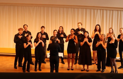 Eine Gruppe junger Sänger*innen steht auf einer Bühne in einem hellen Saal. Sie tragen schwarze Kleidung und singen unter der Leitung eines Dirigenten, die ihnen zugewandt steht. Alle haben die Hände auf der Brust und singen konzentriert.