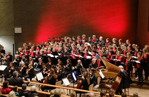 Ein großes Schulorchester und ein Chor treten gemeinsam in einer Kirche auf. Die Musiker*innen spielen Streich- und Blasinstrumente, während der Chor in schwarzen Outfits mit roten Schals singt. Die Bühne ist in warmes rotes Licht getaucht, das Publikum s