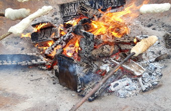 Mehrere Personen halten lange Stöcke mit aufgerolltem Brotteig über ein offenes Lagerfeuer in einer großen Feuerschale. Die Brote backen über den Flammen, im Hintergrund stehen Schüsseln mit Teig.