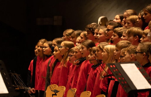 Eine große Gruppe von Kindern steht in Reihen auf der Bühne und singt. Sie tragen rote T-Shirts und blicken aufmerksam nach vorn. Im Vordergrund sind Notenständer zu sehen.