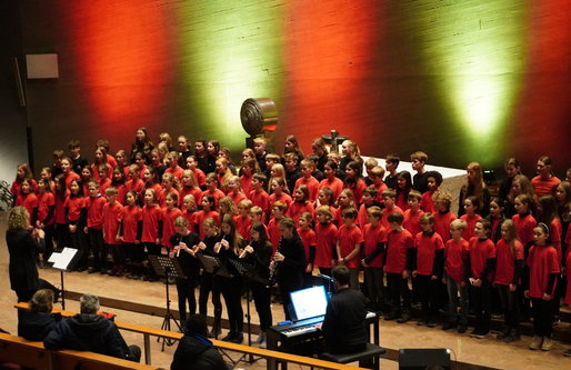 Ein großer Schülerchor mit Kindern in roten T-Shirts singt in einer Kirche. Vor dem Chor spielen einige Schüler*innen Flöte und Oboe, begleitet von einer Person am Klavier. Im Hintergrund ist farbig beleuchtete Kirchenwand in Rot und Grün zu sehen.