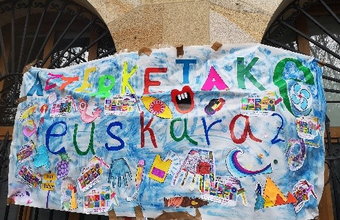 Handgemaltes, farbenfrohes Plakat mit dem Schriftzug „Euskaraz“, gestaltet mit vielen bunten Symbolen, Händen und Zeichnungen. Es hängt an einer Steinwand.