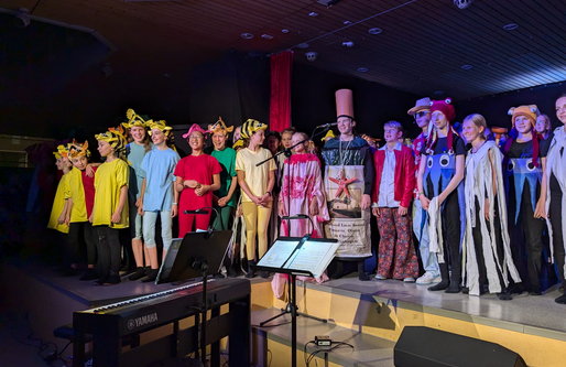 Eine Gruppe von Schüler*innen steht auf einer Bühne in bunten Kostümen und singt. Zu sehen sind fantasievolle Tier- und Meereswesen-Verkleidungen. Vor der Bühne steht ein Klavier mit Notenständern.