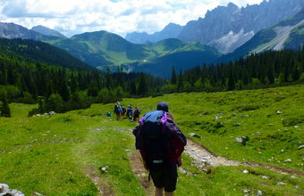 Wanderer mit großen Rucksäcken steigen auf einem schmalen Pfad ins Tal hinab, umgeben von grünen Wiesen, Wäldern und hohen Bergen.