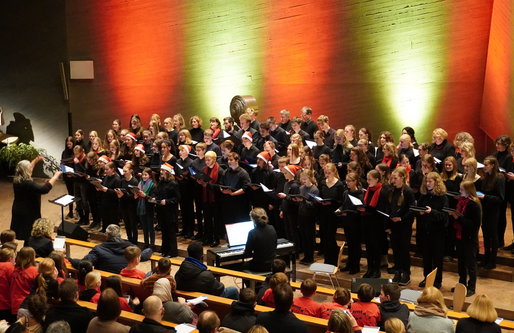 Ein großer Schulchor singt in einer Kirche unter warmem rot-grünem Licht. Die Sänger*innen tragen schwarze Kleidung, einige rote Schals oder Weihnachtsmützen. Eine Dirigentin leitet den Chor, begleitet von einer Person am Klavier. Publikum sitzt in den vo