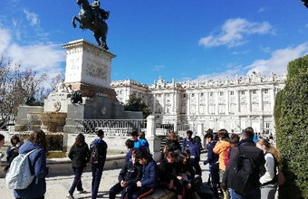 Eine Schüler*innengruppe sitzt und steht bei einer Pause an der Reiterstatue vor dem Königspalast in Madrid. Der Himmel ist blau und sonnig.
