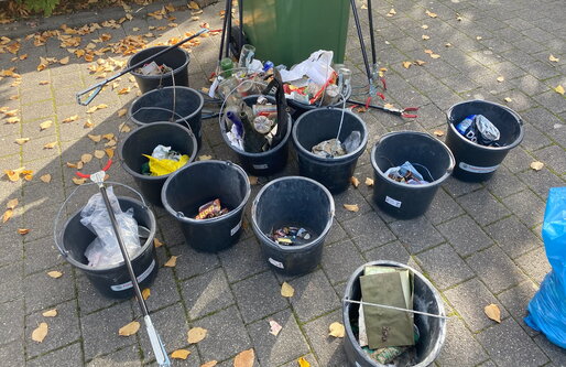 Mehrere schwarze Eimer stehen auf dem Schulhof, gefüllt mit gesammeltem Müll wie Plastik, Papier und Dosen. Daneben liegen Greifzangen – Ergebnis der Müllsammelaktion.