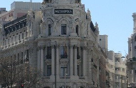 Blick auf das prächtige Metropolis-Gebäude an einer großen Straßenkreuzung in Madrid. Autos fahren vorbei, und die Sonne scheint auf die Fassade mit der schwarzen Kuppel und goldenen Verzierungen.