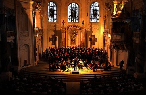 Ein Chor und ein Orchester treten in einer prachtvoll beleuchteten barocken Kirche auf. Die Musiker*innen sitzen im Altarraum, der in warmes goldenes Licht getaucht ist. Das Publikum füllt die Kirchenbänke im Vordergrund.