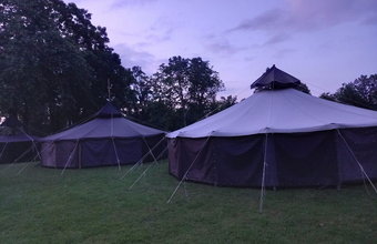 Mehrere große schwarze Jurten stehen auf einer Wiese, umgeben von Bäumen. Der Himmel ist blauviolett in der Abenddämmerung.