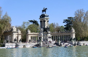 Blick über den See im Retiro-Park in Madrid. Menschen fahren mit Booten, im Hintergrund steht ein Reiterdenkmal mit Säulenbogen und Statue.