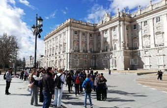 Eine Schüler*innengruppe steht auf dem Platz vor dem Königspalast von Madrid. Das große barocke Gebäude ist in hellem Stein gehalten, der Himmel ist blau mit einigen Wolken.