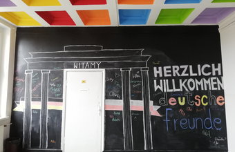 Wandgestaltung mit einer Zeichnung des Brandenburger Tors und der Aufschrift „Witamy“ sowie „Herzlich willkommen deutsche Freunde“. Die Namen der Teilnehmenden sind bunt auf die Tafel geschrieben.