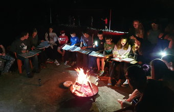 Eine Gruppe von Schüler*innen sitzt abends im Kreis um ein Lagerfeuer. Einige halten Hefte in den Händen und singen, andere leuchten mit Taschenlampen. Das Feuer erhellt ihre Gesichter in der Dunkelheit.