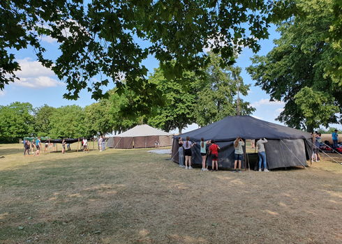 Mehrere Schüler*innen stehen auf einer großen Wiese und bauen Zelte auf. Im Hintergrund sind weitere Zelte und grüne Bäume zu sehen, der Himmel ist blau mit einigen Wolken.