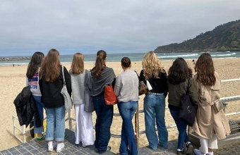 Eine Gruppe Jugendlicher steht mit dem Rücken zur Kamera und blickt auf den Strand und das Meer. Der Himmel ist leicht bewölkt.