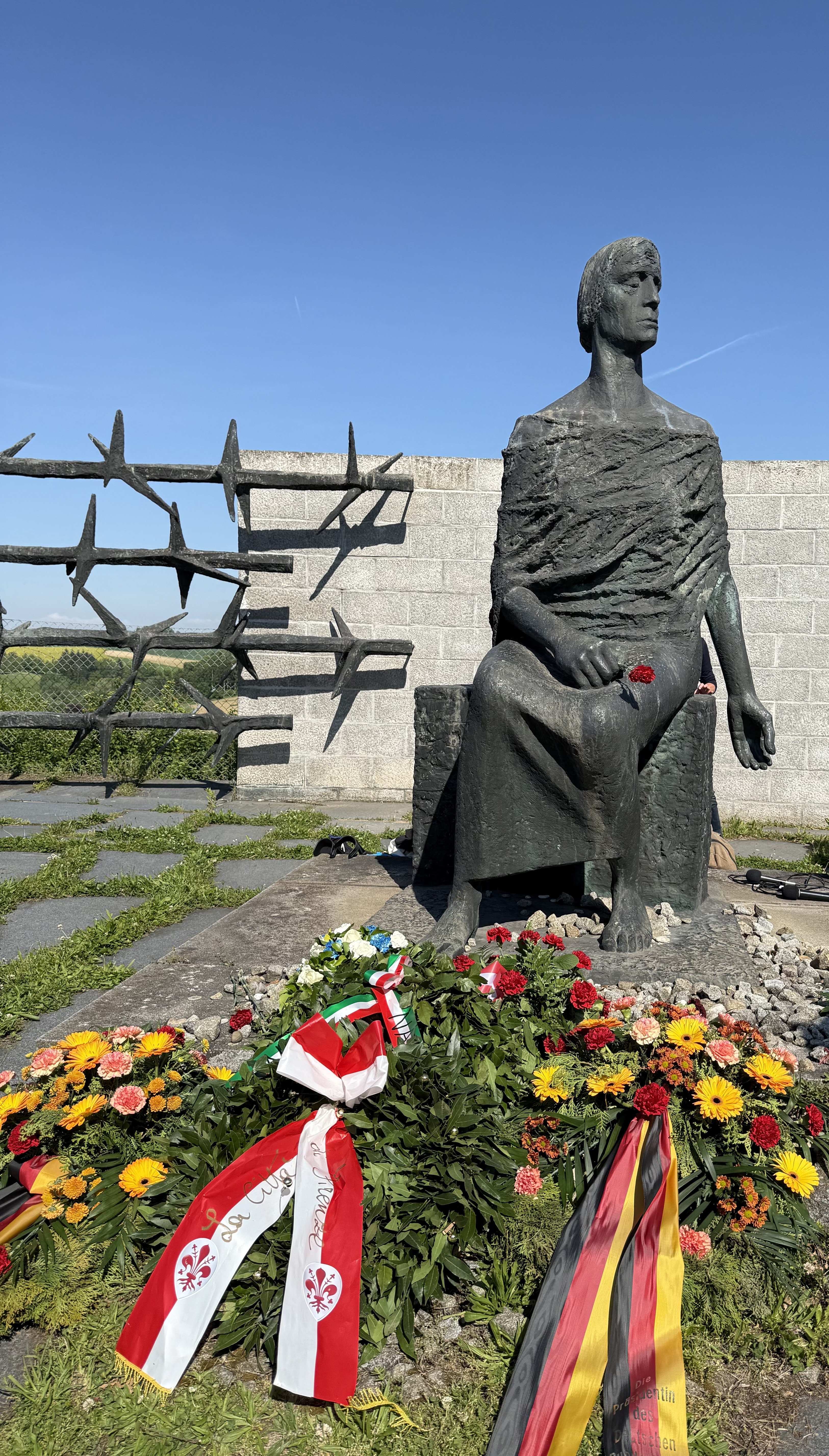 Bronzestatue eines sitzenden Häftlings mit einer roten Blume in der Hand, umgeben von Kränzen und Blumen.